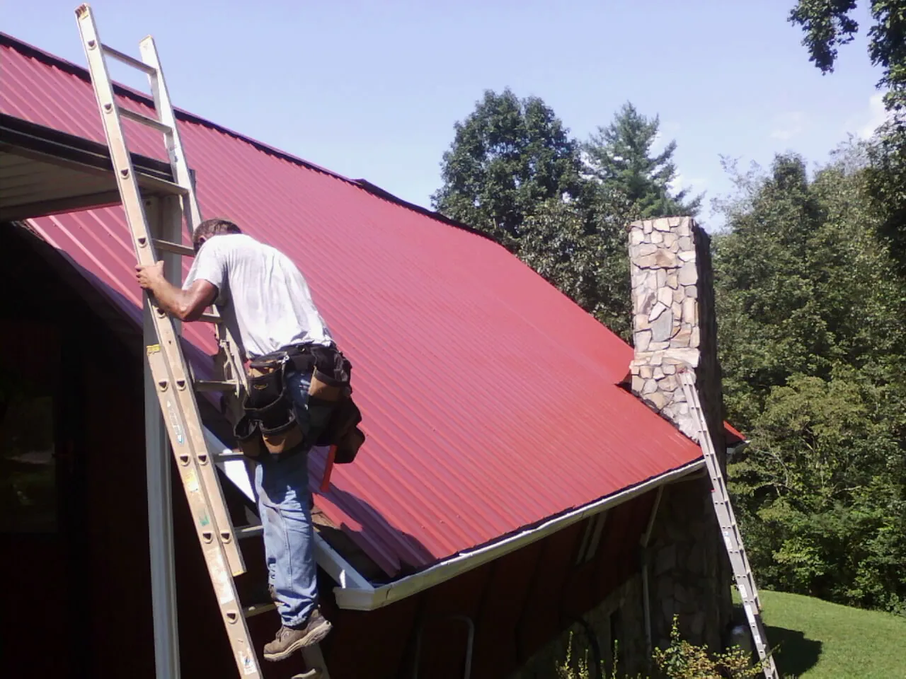 Roofer on a ladder beside a red metal roof and stone chimney — Soffit & Fascia Repair by Roofing Brothers in Eagle Mountain